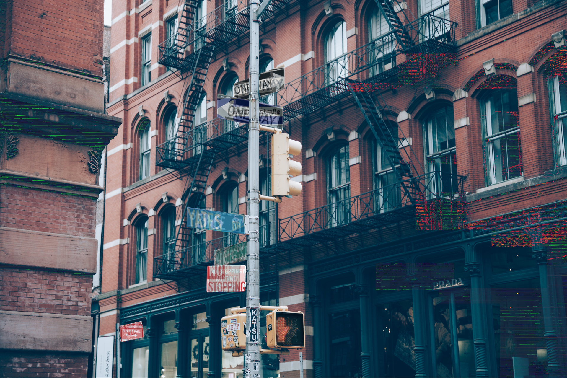 A photo of a city block, with a signpost near the middle, and slight glitch effects around the left and right. The signs hanging from the signpost, as well as another sign in the background, are overlaid with multiple images of the same types of signs.